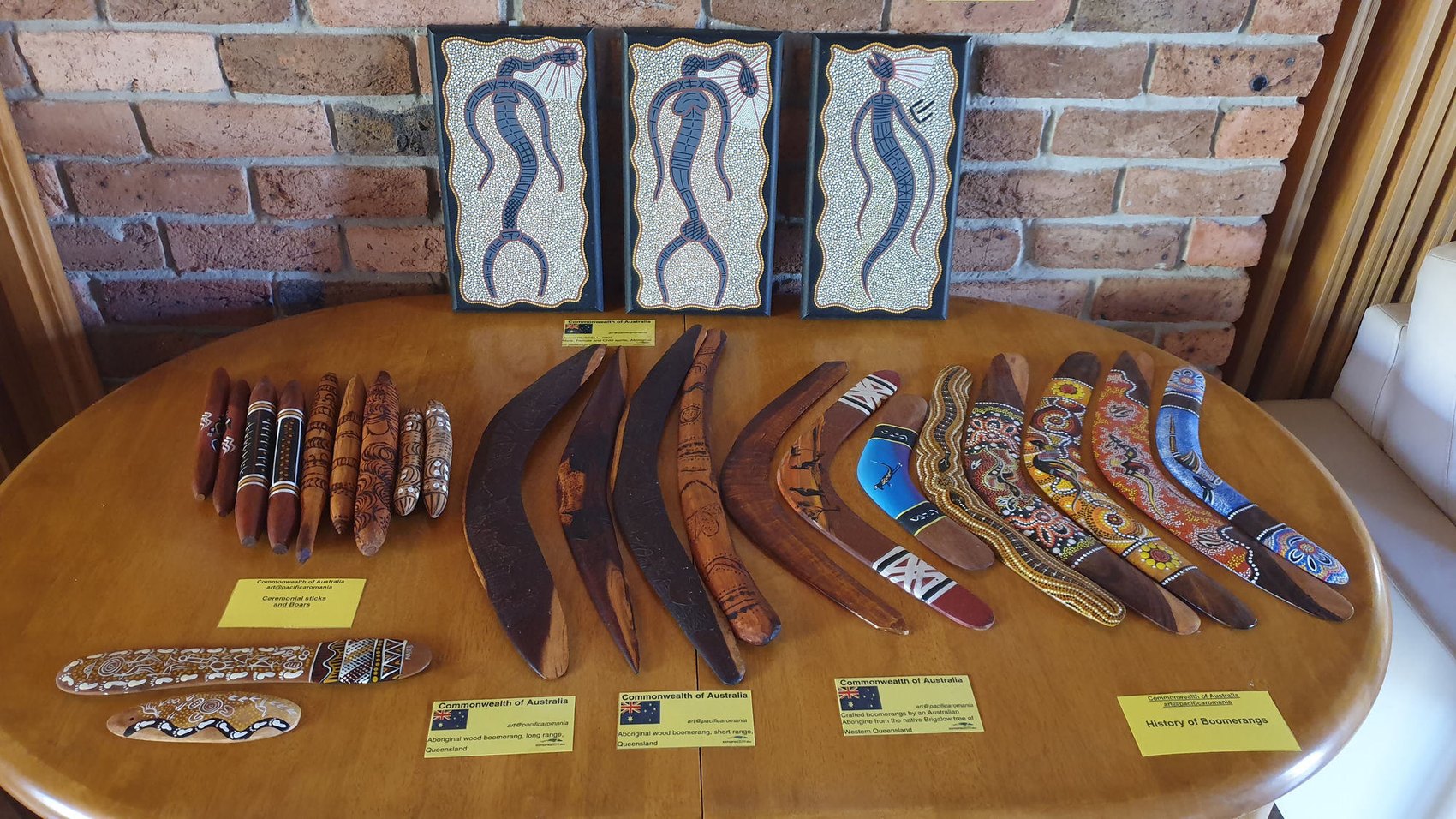 The PacificaRomania boomerang display: three framed Rainbow Serpent panels above ceremonial sticks, incised hunting boomerangs, a pokerwork souvenir and contemporary dot-painted works / Vitrina de bumerange PacificaRomania: trei panouri înrămate cu Șarpele-Curcubeu deasupra unor bețe ceremoniale, bumerange de vânătoare incizate, un suvenir în pirogravură și lucrări contemporane pictate cu puncte
