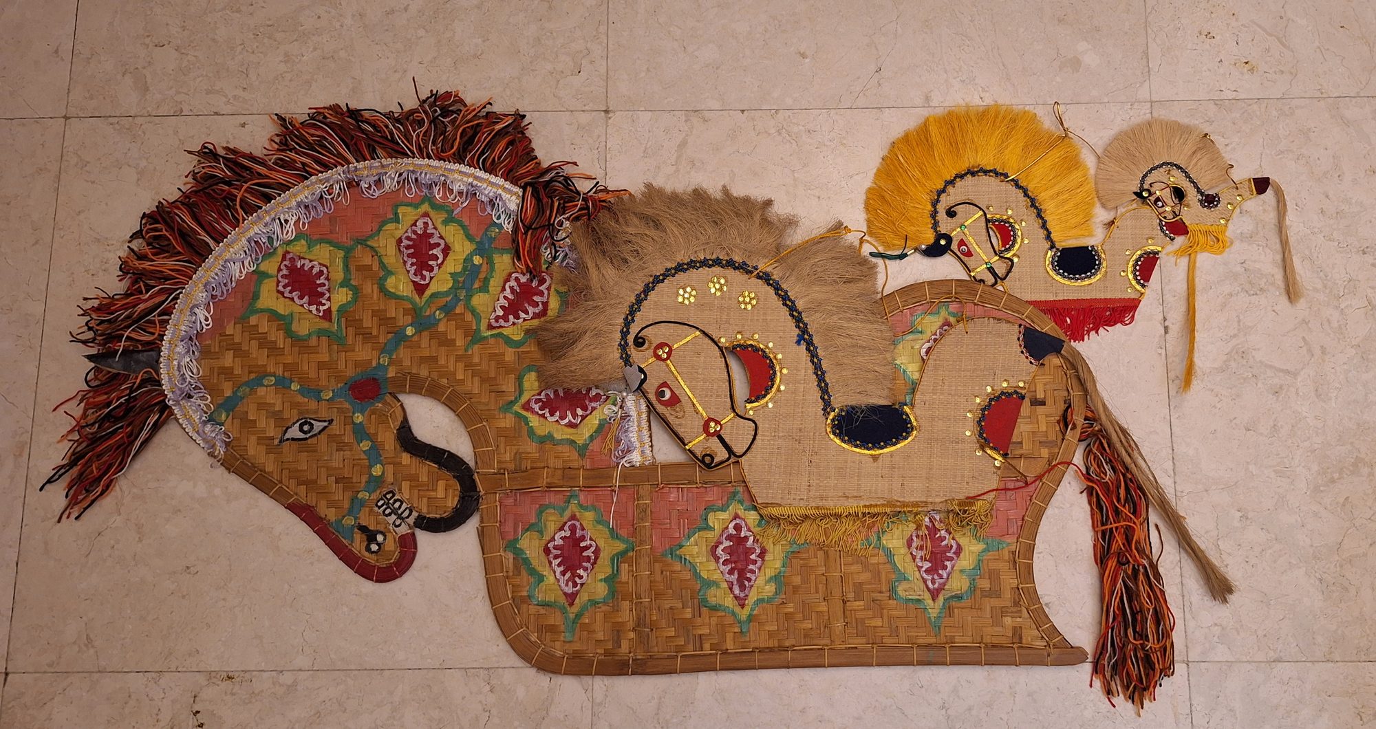 A woven Kuda Kepang horse of plaited bamboo with painted lozenge motifs and a shaggy wool mane, laid beside three smaller gilded, sequinned jute-and-palm-fibre horses with fibre manes / Un cal Kuda Kepang din bambus împletit cu motive romboidale pictate și coamă din lână, alături de trei căluți mai mici din iută și fibră de palmier, auriți și cu paiete