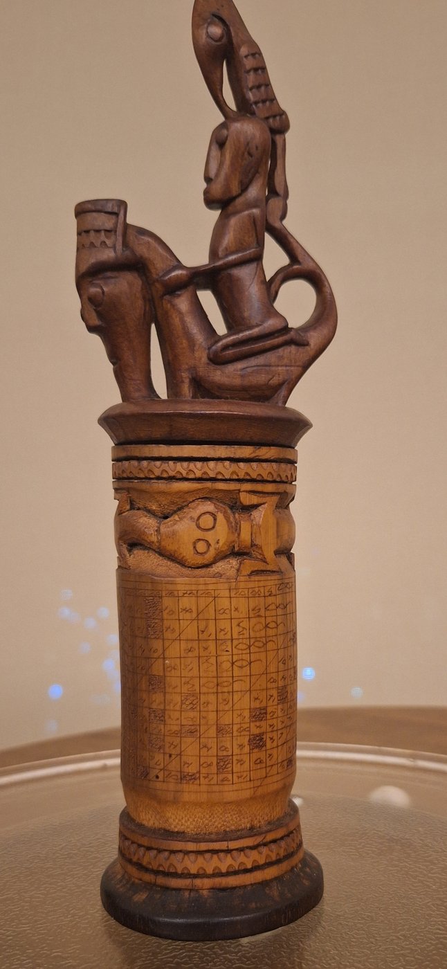Plate 3 — a datu's bamboo container incised with a Batak-script porhalaan calendar grid, topped by a singa-and-rider group
