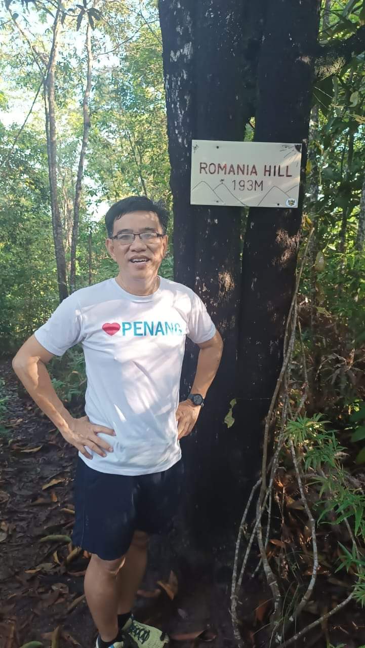 A hiker in an I-love-Penang shirt stands beside a tree on a forested ridge; a hand-lettered white sign nailed to the trunk reads ROMANIA HILL, 193 M / Un drumeț într-un tricou I-love-Penang stă lângă un copac pe o culme împădurită; un semn alb scris de mână, bătut în cuie pe trunchi, spune ROMANIA HILL, 193 M