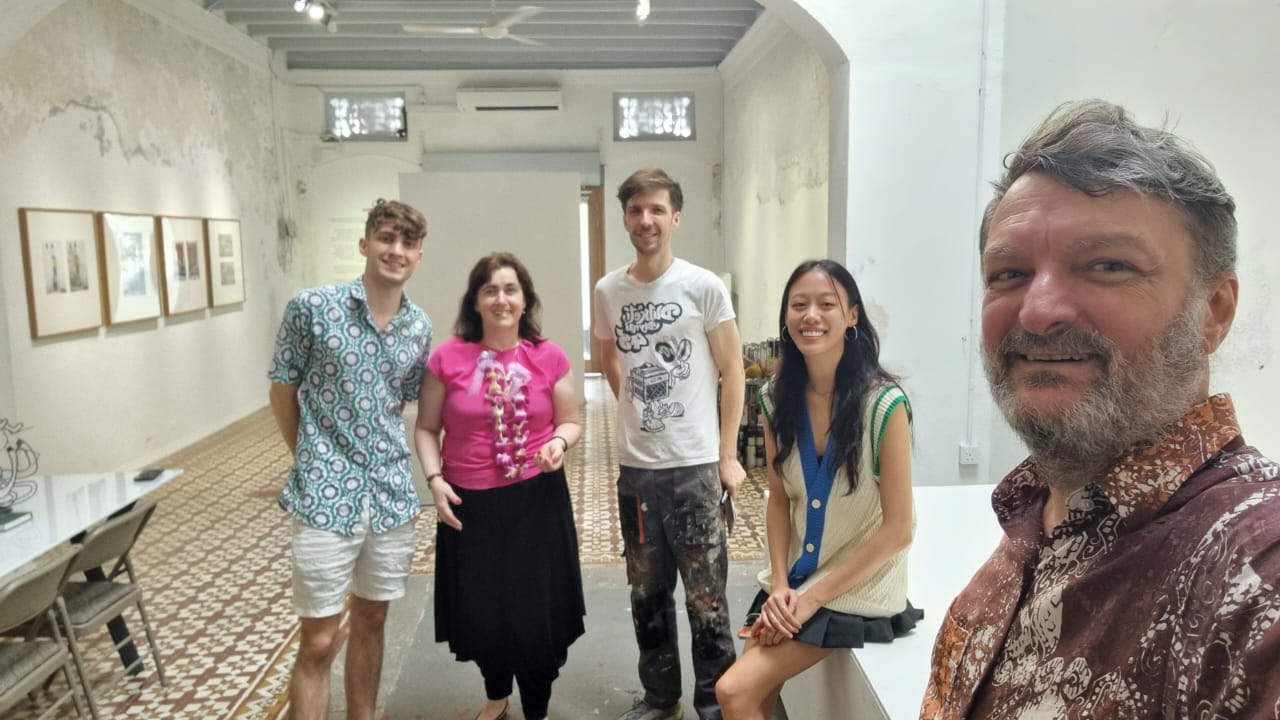 A group portrait in Ernest Zacharevic's George Town gallery-studio: the Barbulescu and Zacharevic families among framed prints on a distressed-plaster wall and a patterned encaustic-tile floor, the artist in the foreground / Un portret de grup în atelierul-galerie al lui Ernest Zacharevic din George Town: familiile Bărbulescu și Zacharevic între tipărituri înrămate pe un perete de tencuială scorojită și o podea cu plăci decorate, artistul în prim-plan