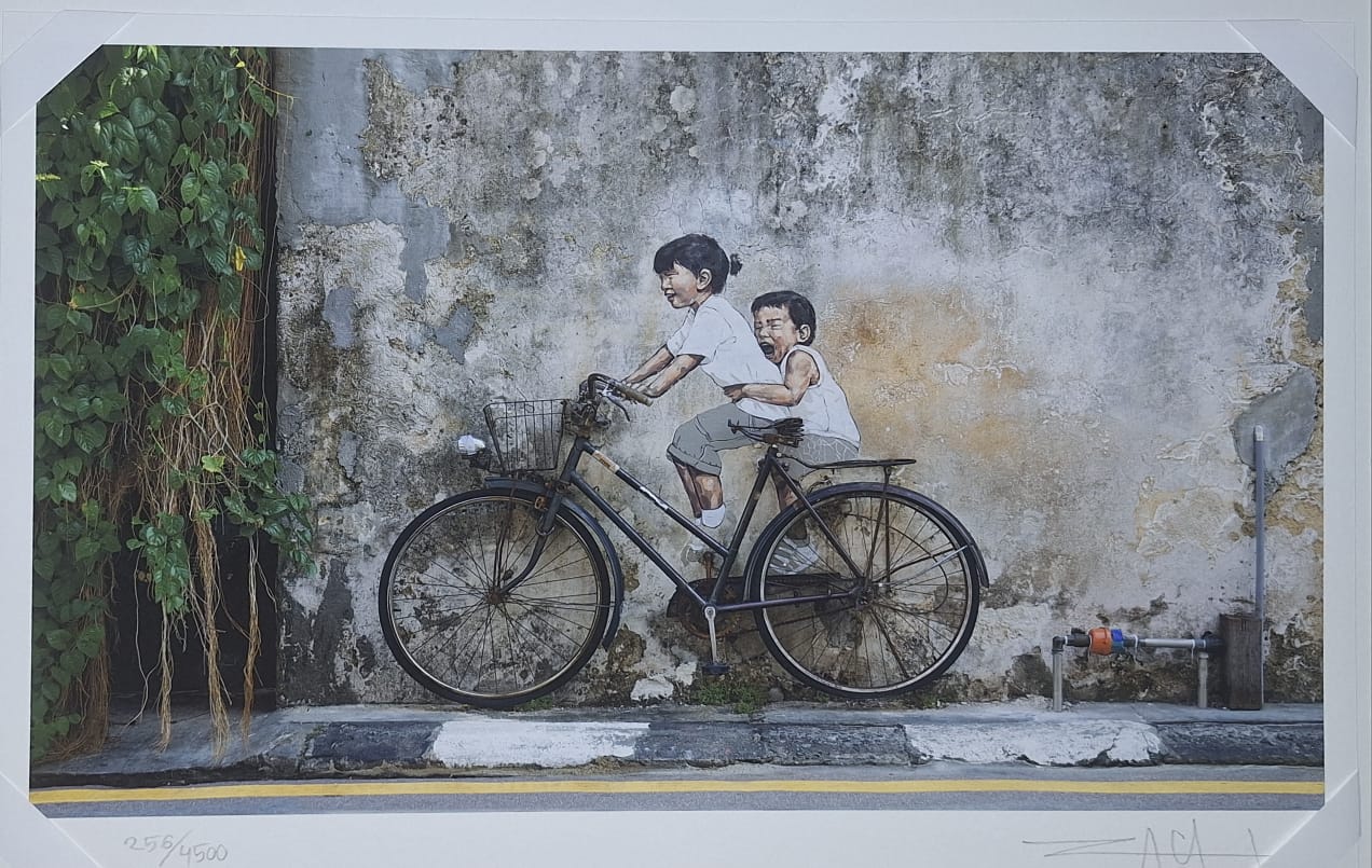 Signed limited-edition print of Ernest Zacharevic's mural Little Children on a Bicycle: two children on a real bicycle painted against a weathered George Town wall, a trailing vine at left and a yellow road line below / Tipăritură semnată, în ediție limitată, a muralei Micii copii pe bicicletă de Ernest Zacharevic