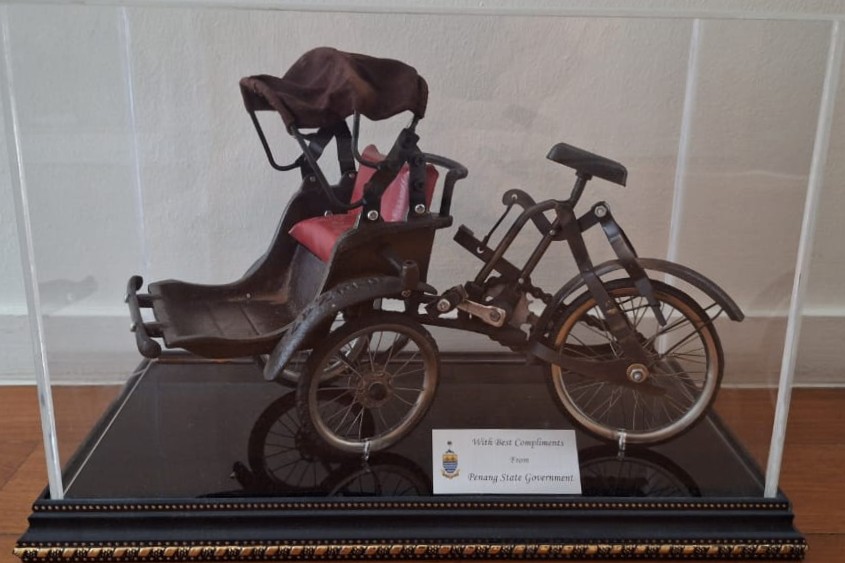 A bronze-finished model of a Penang trishaw (beca) with a red passenger cushion in a clear acrylic display case, set before the framed print of the bicycle mural; a plaque reads With best compliments from Penang State Government / Un model cu finisaj de bronz al unui trishaw din Penang (beca), cu pernă roșie, într-o casetă de acril, în fața tipăriturii înrămate a muralei cu bicicleta