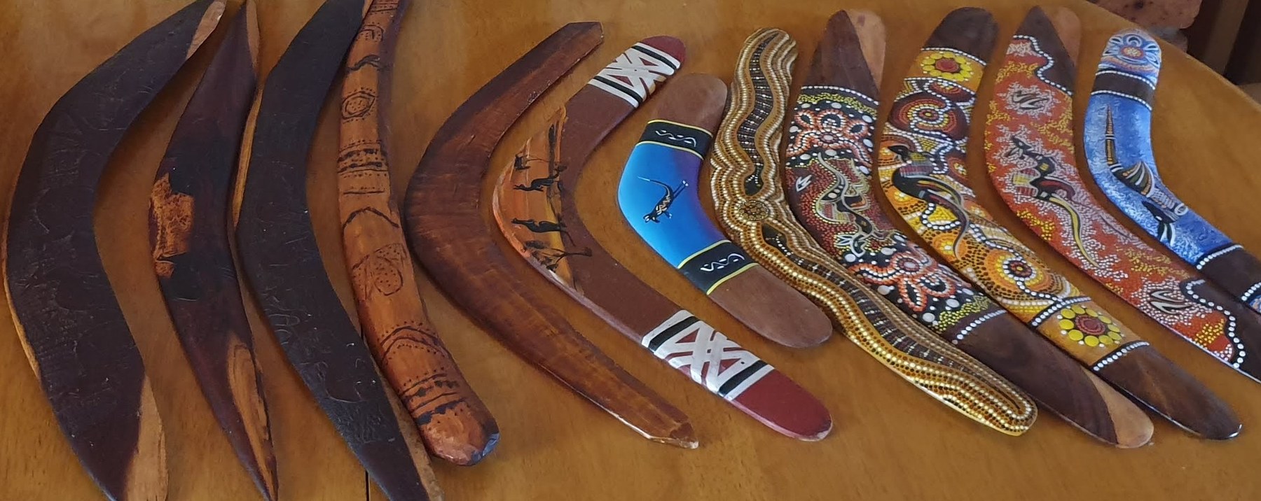 A row of Aboriginal boomerangs on a honey-wood table, from dark incised hardwood hunting forms to vibrantly dot-painted and pyrography-decorated contemporary pieces / Un șir de bumerange aborigene pe o masă de lemn de miere, de la forme de vânătoare din lemn de esență tare incizat la piese contemporane pictate cu puncte și decorate în pirogravură